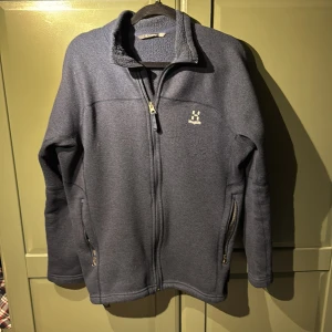 Mörkblå fleecejacka från Haglöfs - Mysig mörkblå fleecejacka från Haglöfs med hel dragkedja och höga fickor med dragkedja. Jackan har ståkrage och är perfekt för kyliga dagar. Klassisk och enkel design med diskret logga på bröstet. Passar dig som gillar outdoor och chill vibes.