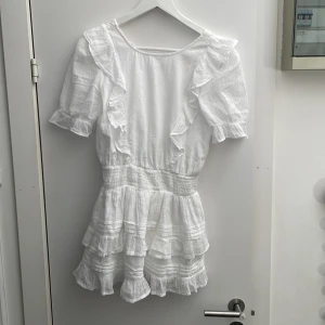 Vit volangklänning med kort ärm🌸 - Superfin vit klänning med volanger och kort ärm. Klänningen har v-ringning med knyt i nacken och smock i midjan för en markerad siluett. Kjolen är kort med flera lager volanger för extra volym. Perfekt för sommarens alla tillfällen!🩷