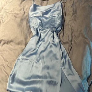 Ljusblå långklänning i satin - Supersnygg ljusblå långklänning i glansigt satinliknande material med tunna axelband och figurnära passform. Klänningen har en slits framtill och en elegant, enkel design som ger en lyxig känsla. Perfekt för dig som vill sticka ut med stil. Har två små fläckar i fram som knappt syns men som jag ska försöka få bort i tvätten.