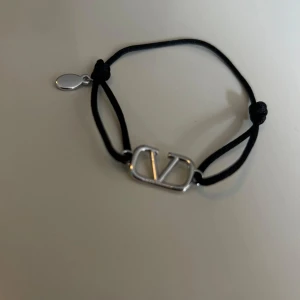 Valentino svart armband med silverdetalj - Snyggt armband från Valentino med svart justerbart band och en stilren silverfärgad logga i metall. Perfekt accessoar för att lyfta din outfit med en lyxig touch. Diskret bricka med Valentino-gravyr vid knuten. Kvitto från Farfetch tillgängligt
