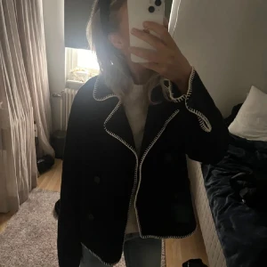 Svart skepparkavaj med kontrastsömmar - Säljer en svart skepparkavaj med vita kontrastsömmar längs krage, fickor och ärmslut. Jackan har dubbelknäppning, bred krage och raka fickor framtill. Perfekt för dig som gillar en stilren men ändå unik look.