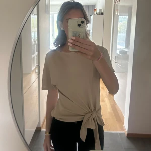 Beige topp med knyt i sidan - Säljer en beige topp med korta ärmar och snygg knytning i sidan. Enkel och stilren design med rund halsringning, perfekt att matcha med jeans eller kjol.
