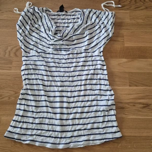 Randig vit och blå linnetopp - Snygg vit linnetopp med marinblå ränder och dragsko vid axlarna. Toppen har en lös passform och är perfekt för varma dagar. Tunt och mjukt material som känns skönt mot huden.