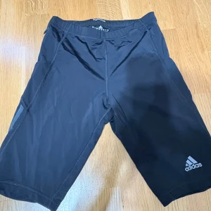 Svarta träningsshorts från Adidas - Svarta Adidas shorts med klassiska grå ränder på sidorna och logga framtill. De har elastisk midja med snörning och är gjorda i lätt syntetmaterial som andas. Perfekta för sport och träning, med normal passform och knälång modell.