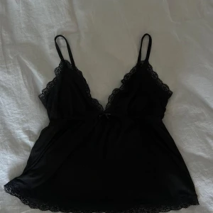 Top från Nelly - Säljer en svart linnetopp med tunna axelband och v-ringning från Nelly. Toppen har fina spetsdetaljer längs urringningen och nederkanten samt en liten rosett framtill.
