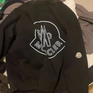 Svart Moncler sweatshirt med stor logga - Svart sweatshirt från Moncler med stor vit logga på ryggen och klassisk Moncler-patch på ärmen. Tröjan har en avslappnad passform och är tillverkad i mjuk bomull, perfekt för en stilren och trendig look.