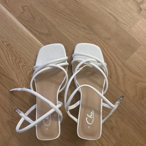 Vita sandaler med klack från Color - Snygga vita sandaler med fyrkantig tå och smala remmar som korsar foten. Skorna har en låg klack och spänne vid ankeln. Dom är använda 1 gång! 
