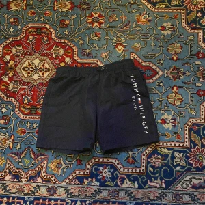 Blå badshorts Tommy Hilfiger - Snygga marinblå badshorts från Tommy Hilfiger med vit logga längs ena benet. Shortsen har elastisk midja och innerbyxa i mesh för extra komfort. Perfekta för stranden eller poolen, klassisk stil med sportig vibe.