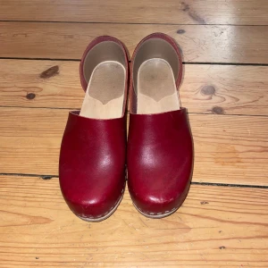 Röda träskor med klack - Säljer ett par klassiska röda träskor i skinn med träklack och platt sula. Skorna har en enkel och stilren design med öppen häl och rund tå. Perfekta för dig som gillar en retrovibe och vill ha något som sticker ut. Storlek 35.