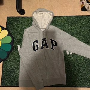 Grå hoodie-dragkedja från GAP - Snygg grå hoodie från GAP med stor broderad logga i svart och vitt på bröstet. Tröjan har huva med snörning, dragkedja framtill och två fickor. Tillverkad i mjuk bomullsmix.