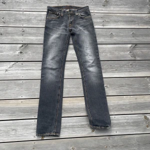 Svarta Nudie Jeans - Snygga svarta jeans från Nudie Jeans med straight fit och slitna detaljer. Jeansen har orange kontrastsömmar och ikonisk brodering på bakfickorna. Tillverkade i mjuk bomullsdenim med dragkedja och knapp framtill. Jeansen ganska low waist, och helt oanvända.