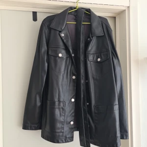 Svart skinnjacka från H&M Divided S - Snygg svart tunn skinnjacka från H&M Divided i storlek S. Jackan har klassisk krage, silverfärgade knappar, två bröstfickor med lock och två stora fickor framtill. Rak passform och långa ärmar, perfekt för en edgy look.