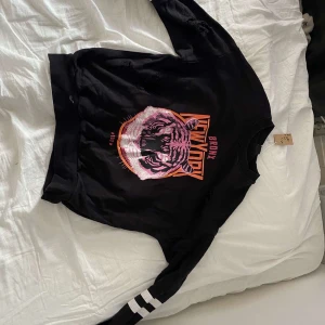 Svart sweatshirt med tigertryck Gina Tricot - Cool svart sweatshirt från Gina Tricot med stort tigertryck i rosa och orange samt texten 'Bronx New York'. Tröjan har vita ränder på ärmarna och slitna detaljer för en edgy look. Perfekt för dig som gillar streetstyle. Obs! hålen är design