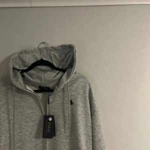 Grå hoodie från Polo Ralph Lauren - Snygg grå hoodie från Polo Ralph Lauren med dragkedja och huva. Klassisk broderad logga på bröstet i mörkblått. Tröjan har snörning i huvan och ribbade muddar. Perfekt för en chill och stilren look.