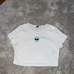 Vit croppad t-shirt med alientryck - Cool vit croppad t-shirt från Divided med ett mintgrönt alientryck på bröstet. T-shirten har rund hals och korta ärmar, perfekt för dig som gillar streetstyle och vill sticka ut med en unik detalj. Materialet är mjuk bomull som känns skönt mot huden.