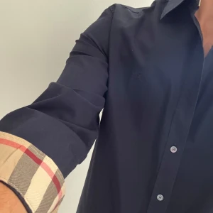 Mörkblå Burberry skjorta med rutig detalj - Snygg mörkblå skjorta från Burberry med klassisk rutig insida på krage och manschetter. Skjortan har knappar framtill, lång ärm och diskret Burberry-logga broderad på bröstet. Perfekt för dig som gillar stilrena plagg med exklusiva detaljer. Passar medium perfekt! 