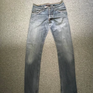 Nudie Jeans blå slim fit jeans - Storlek: 30/32.       Blå jeans från Nudie Jeans med klassisk femficksdesign och snygga slitningar. Jeansen har raka ben, orangea sömmar och Nudie-logga på bakfickan. Tillverkade i slitstarkt bomullsjeans med en cool tvättad look. 