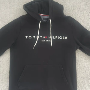 Svart hoodie från Tommy Hilfiger M - Svart hoodie från Tommy Hilfiger med vit logga och text på bröstet. Klassisk huva med vita snören och stor magficka framtill. Perfekt för chill dagar och enkel att matcha med jeans eller joggers. Använd 3 gånger! 