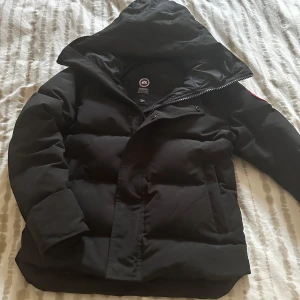 Svart jacka från Canada Goose - Säljer en svart jacka från Canada Goose med huva och klassisk logga på ärmen. Jackan har dragkedja och knappar framtill samt innerficka med dragkedja. Perfekt för kalla dagar och riktigt snygg streetstil. PRIS KAN DISKUTERAS!!