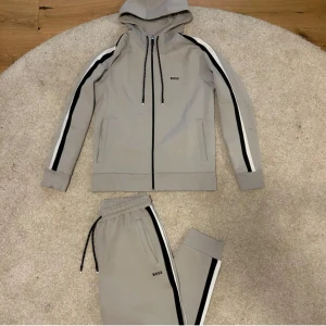 Beige BOSS tracksuit - Snyggt beige tracksuit från BOSS i storlek M. Setet består av en hoodie med dragkedja, svarta och vita ränder längs ärmar och ben, samt svarta snören med vita detaljer. Byxorna har elastisk midja med snörning och sidofickor. Perfekt för chill eller träning.