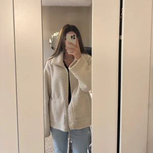 Beige teddyjacka  - Mysig beige teddyjacka med dragkedja framtill och hög krage. Jackan har en loose passform och är perfekt för kyliga dagar. Tillverkad i fluffigt fleece-material som ger en skön och avslappnad vibe.