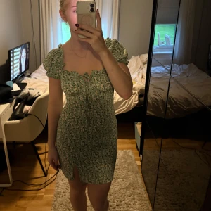 Grön blommig kortklänning med puffärm - Supersöt grön kortklänning med små vita blommor, puffärmar och söt knytning vid bysten. Klänningen har en liten slits framtill och figurnära passform. Perfekt för sommarens alla tillfällen och ger en romantisk vibe.