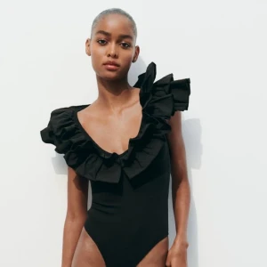Body från Zara med volanger  - Supersnygg svart body med breda volanger runt axlar och halsringning. Modellen har djup v-ringad fram och öppen rygg, vilket ger en riktigt cool och trendig look. 