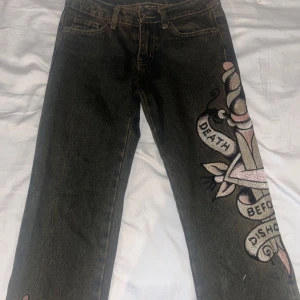 Ed Hardy jeans med broderi, 26W - Snygga svarta jeans från Ed Hardy med stort broderat motiv på benet i rosa, silver och beige. Klassisk femficksmodell med Ed Hardy-logga på bakfickan och coolt tattoo-inspirerat mönster. Raka ben och normal passform, perfekt för dig som gillar statement-plagg.