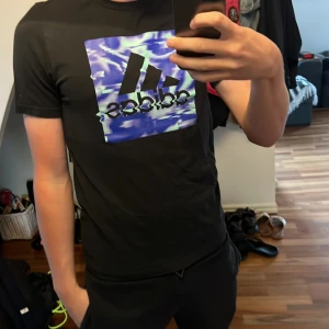 Svart Adidas t-shirt med blått tryck - Svart t-shirt från Adidas med ett stort, färgstarkt tryck på bröstet i blått och lila toner. Trycket har en cool, vågig effekt och den klassiska Adidas-loggan. T-shirten har rund hals och korta ärmar, perfekt för en sportig och chill stil.