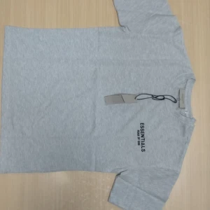 Grå Essentials Fear of God t-shirt - Snygg grå t-shirt från Fear of God Essentials med svart tryck på bröstet och ryggen. Klassisk rund hals och korta ärmar. Tillverkad i mjuk bomull som känns skön mot huden. Perfekt för dig som gillar streetwear och cleana vibes. Nypris runt 1200 kr. Sprillans ny kommer med lappar och påse. Tröjan för 499 kr. Om du även vill ha shortsen så kan du få båda för 799.