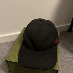 Svart keps från Gucci med GG-mönster - Snygg svart keps från Gucci med diskret GG-monogram över hela tyget. Klassisk form med böjd skärm och justerbar rem bak med grönt och rött band. Kepsen är tillverkad i bomull och polyester, perfekt för dig som gillar exklusiva accessoarer. Den är även äkta.