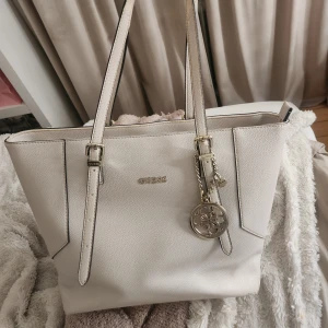 Beige väska från Guess - Snygg beige stor väska från Guess med justerbara remmar och guldfärgad logga samt hänge. Väskan är i strukturerat skinnimitation. Perfekt storlek för att få plats med allt du behöver och ger en clean look.