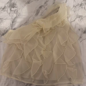Champagnefärgad omlottklänning med volanger - Superfin kort klänning i champagnefärg med ett axelbandslöst one-shoulder snitt. Klänningen har flera lager av tunna volanger som ger en romantisk och luftig känsla. Perfekt för dig som vill sticka ut med en elegant och unik look.