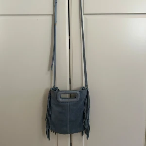 Blå mocka axelväska med fransar från maje  - Supersnygg blå axelväska i mocka från maje med långa fransar på sidorna och både axelrem och handtag. Perfekt storlek för det viktigaste och ger en cool boho-vibe till din outfit.
