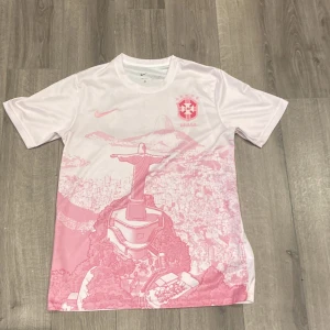 Vit och rosa Brasilien fotbollströja Nike - Snygg Brasilien fotbollströja från Nike i vitt och rosa med ikoniskt tryck av Kristusstatyn och Rio de Janeiro. Tröjan har korta ärmar, rund hals och är gjord i ett lätt och ventilerande material som passar perfekt till träning eller match.