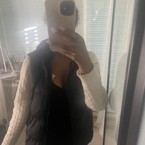 Svart dunväst med dragkedja - Säljer en svart dunväst med hög krage och dragkedja framtill. Västen har en enkel och stilren design som passar perfekt över en stickad tröja. Den är lätt vadderad och har en klassisk passform.