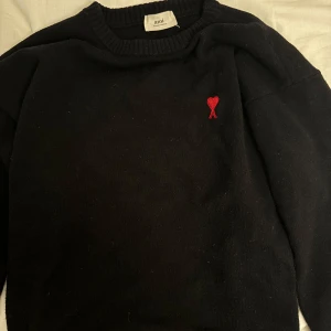 Svart stickad tröja från Ami Paris - Svart stickad tröja från Ami Paris med röd broderad logga i form av ett hjärta på bröstet. Klassisk rund halsringning och ribbade muddar. Perfekt för dig som gillar stilrena plagg med en twist.