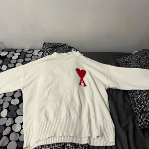 Vit stickad tröja från Ami Paris - Vit stickad tröja från Ami Paris med röd hjärtlogga och bokstaven A på bröstet. Tröjan har ribbade muddar och krage, samt en relaxed passform. Perfekt för dig som vill ha en stilren men ändå unik look.