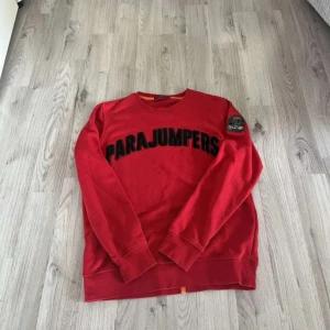 Röd Parajumpers sweatshirt i bomull - Röd sweatshirt från Parajumpers med stort svart broderat logotyptryck över bröstet och patch på ena ärmen. Tröjan är i 100% bomull och har rund hals samt långa ärmar. Perfekt för dig som gillar streetwear och vill sticka ut.