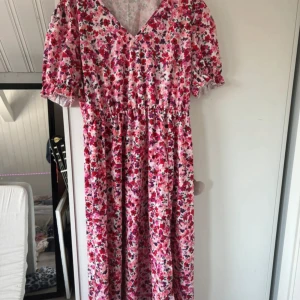 Blommig långklänning med puffärm - Säljer en superfin långklänning med blommigt mönster i rosa, rött och lila toner. Klänningen har v-ringning, puffiga kortärmar med resår och volangdetalj samt markerad midja. Materialet är mjukt och lätt, perfekt för varma dagar. Så söt och färgglad!