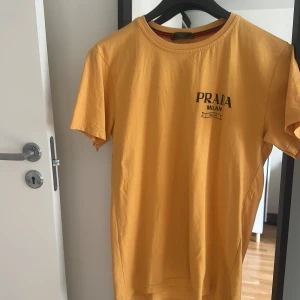 Gul t-shirt från Prada Milano - Gul t-shirt från Prada Milano med svart tryck både fram och bak. Klassisk rund hals och korta ärmar. Materialet är mjuk bomull och passformen är normal. Perfekt för dig som gillar stilrena designer och vill sticka ut med en ikonisk logga.