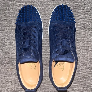 Mörkblå skor från Christian Louboutin - Säljer ett par mörkblå/marinblå skor från Christian Louboutin med coola spikar/nitar på tån och klassisk röd sula. Skorna har snörning och är gjorda i mocka med vita platta sulor. Insidan är beige och de har en rund tå. Perfekt för dig som vill sticka ut med lyxig streetstyle. 