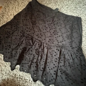 Svart volangkjol med broderade detaljer - Supersöt svart kortkjol från Lindex med broderade blommor och hålmönster. Kjolen har volangkant och vågig nederdel som ger en extra touch. Perfekt för dig som gillar feminina detaljer och vill sticka ut lite extra.