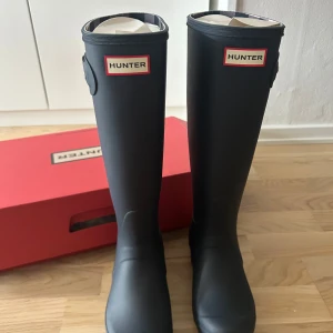 Mörkblå Hunter stövlar med rutigt mönster - Snygga oanvända mörkblåa Hunter stövlar i gummi med rutigt mönster baktill. Klassisk hög modell med rund tå och platt sula. Kommer i originalkartong.
