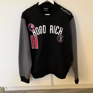Svart och grå bomberjacka från Hoodrich - Säljer en svart och grå bomberjacka från Hoodrich med coola patches och broderade detaljer framtill. Jackan har tryckknappar, ribbade muddar och kontrastärmar i grått. Perfekt för dig som vill sticka ut med streetstyle och sportig vibe. Tröjan är bara använd en få antal gånger och är därför i bra skick!