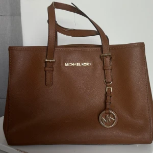 Brun handväska från Michael Kors - Snygg brun handväska från Michael Kors i klassisk boxig modell. Väskan är gjord i strukturerat skinn och har guldfärgade metalldetaljer samt MK-hänge. Två rejäla handtag och justerbara remmar gör den både praktisk och stilren. Den köptes på Jackie för ca 3000+ kr. Den är ganska så använd och har slitningar så därför ger jag den för 800 kr!❤️ 