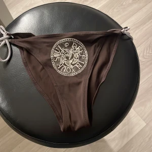 Bruna bikiniunderdel från Skinny - Snygg brun bikiniunderdel från Skinny i storlek 38 med vita knytband i sidorna. Framsidan har ett coolt, cirkulärt tryck i vitt med grafiskt motiv. Materialet är mjukt och stretchigt, perfekt för strandhäng och poolparty.