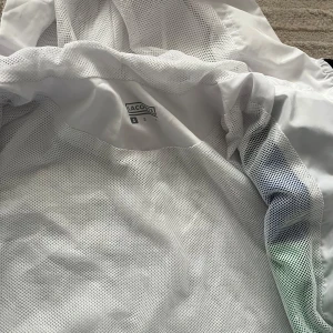 Lacoste vindjacka med färgblock - Snygg vindjacka från Lacoste i vitt med blåa och gröna färgblock samt svarta detaljer. Jackan har huva, dragkedja framtill och meshfoder på insidan. Klassisk krokodil-logga på bröstet och två fickor med dragkedja. Perfekt för dig som gillar sportig och fräsch stil.