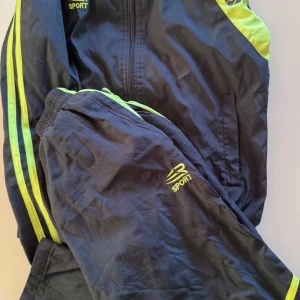 Svart och neongul vindjacka DR Sport - Sportig svart vindjacka från DR Sport med neongula detaljer och dragkedja framtill. Jackan har två fickor och coola ränder längs ärmarna. Perfekt för barn för träning eller andra aktiviteter då de är väldigt bekväma och sköna att ha 