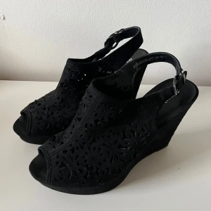 Svarta kilklackar från Graceland - Svarta sandaler med kilklack från Graceland. Skorna har snyggt utskuret blommönster i mockaimitation, öppen tå och justerbar rem med spänne runt hälen. Klackhöjd ungefär 12cm.